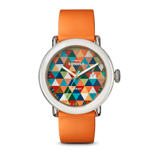 A Shinola Detroit analog watch with a round silver-tone case, a vibrant multicolor geometric triangle dial, and a bright orange strap, shown against a plain white background.